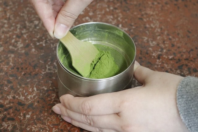 Sifting matcha powder through a fine mesh sieve with a bamboo scoop