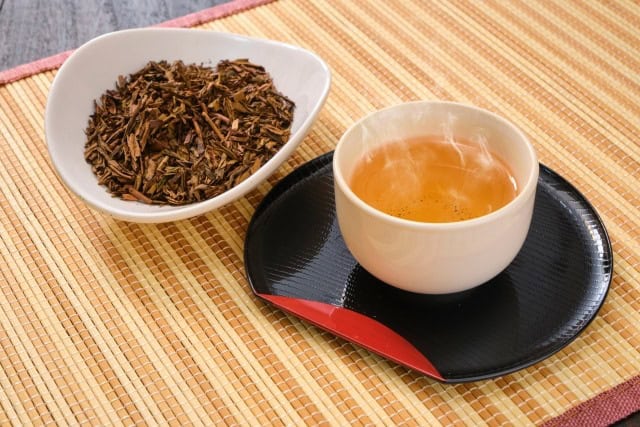 Hojicha tea steaming in a cup beside a bowl of roasted tea leaves