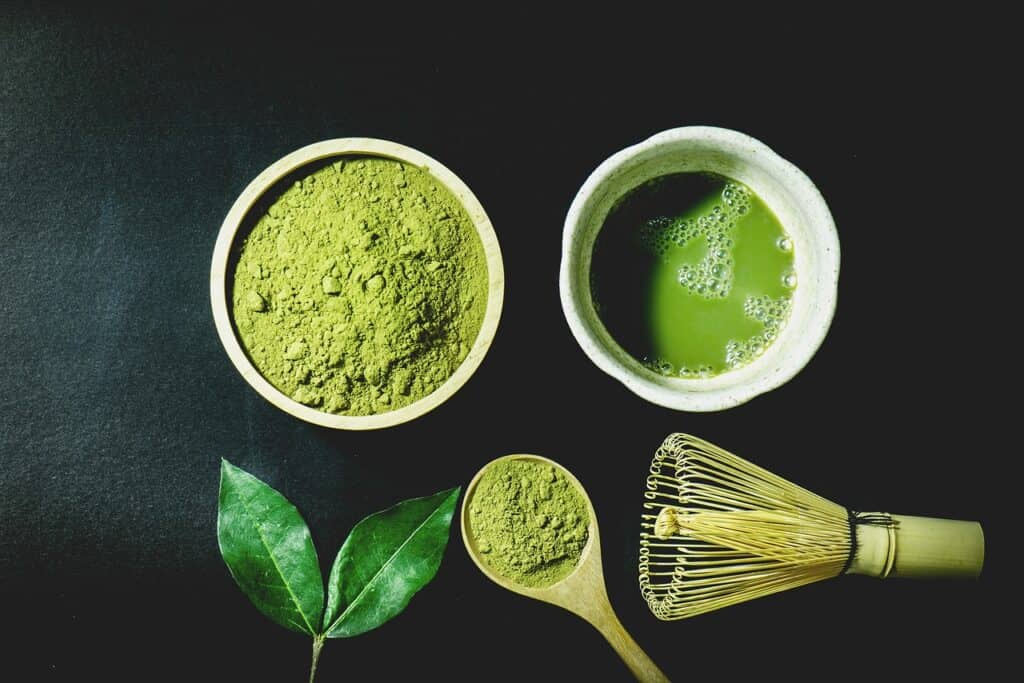 Matcha powder, a bowl of tea and a bamboo whisk on a dark surface